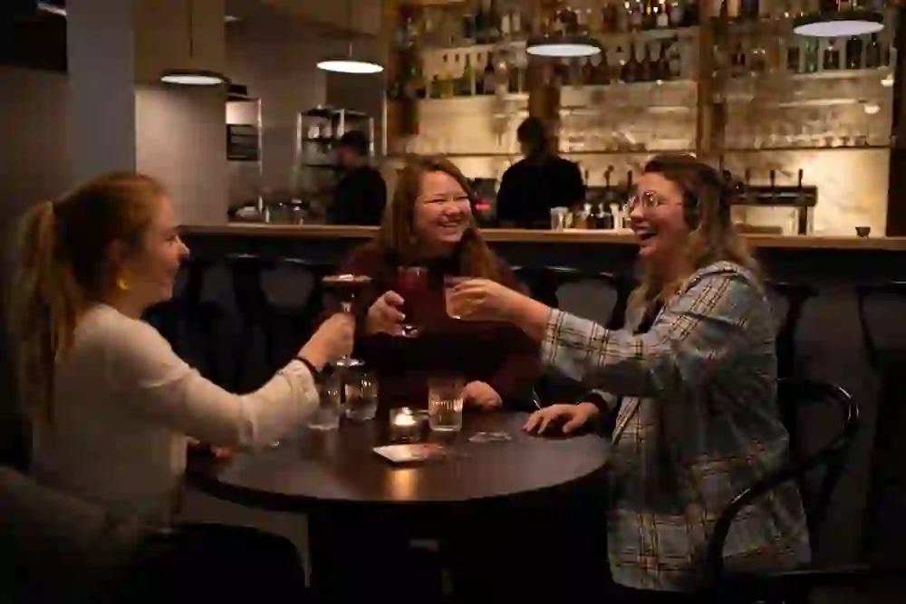 Das Bild zeigt drei Frauen in einer Bar, die bei warmem Licht lachen und miteinander anstoßen. Die entspannte und fröhliche Stimmung ist ansteckend und lädt ein, sich dem Vergnügen anzuschließen.