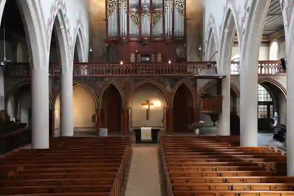 L'image montre l'intérieur de l'église française de Berne à l'architecture gothique. On y voit de longues rangées de bancs en bois qui convergent vers une zone d'autel éclairée avec une grande croix. Au-dessus se trouve un orgue impressionnant sur une tribune, et les murs sont ornés de peintures et de motifs décoratifs. L'ensemble dégage une atmosphère solennelle et historique.