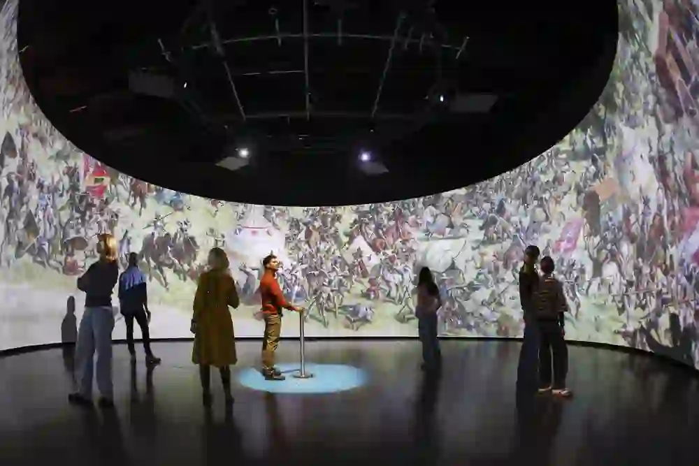 In einem dunklen Raum stehen Besuchende vor einer monumentalen Rundprojektion der Schlacht von Murten. Das detailreiche Panorama umgibt sie vollständig und zieht sie ins Geschehen. Die Stimmung ist eindringlich, nachdenklich und von historischer Wucht geprägt.