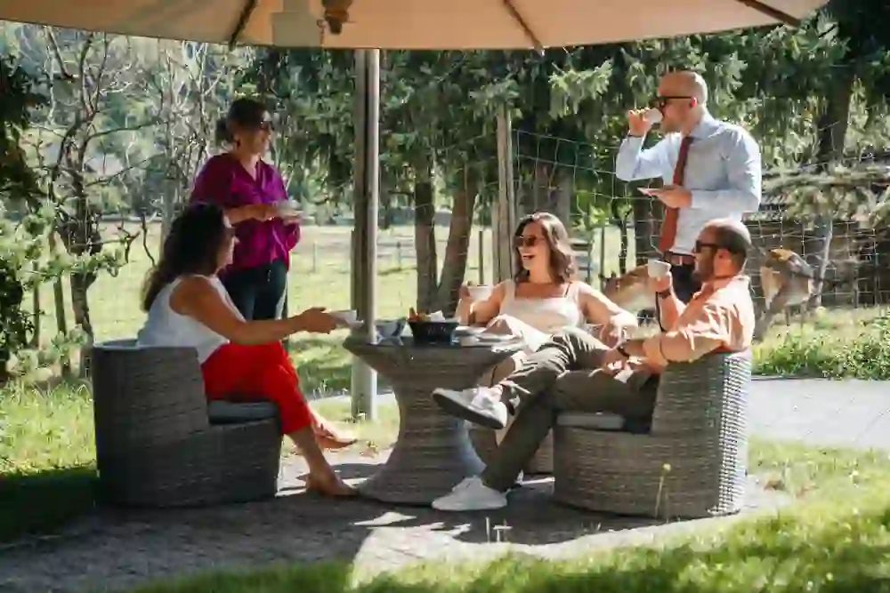 La photo montre cinq personnes assises en toute décontraction sur une terrasse, sous un parasol, en train de boire un café. L'ambiance est détendue et conviviale, ils rient et profitent de la nature dans un cadre idyllique et verdoyant.