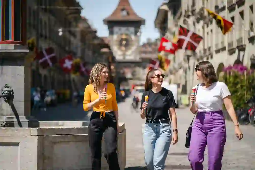 Three people stroll through a busy street in Bern, laughing with ice cream, past historic buildings and Swiss flags - a picture of carefree summer enjoyment.