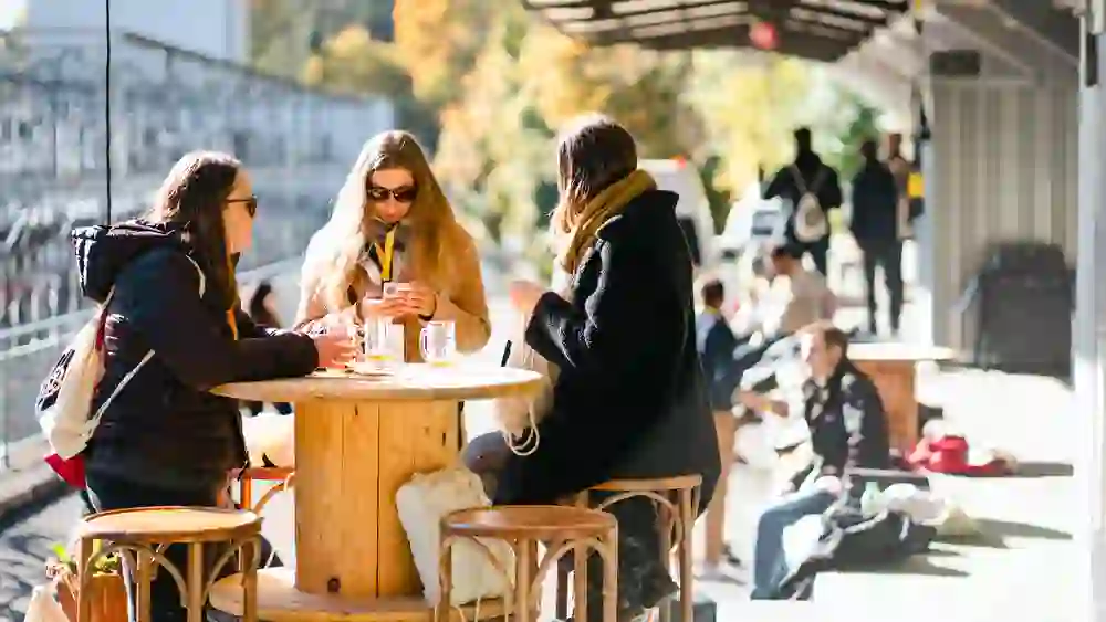 Das Bild zeigt drei Frauen, die um einen runden Holztisch im Freien sitzen. Sie tragen Sonnenbrillen und warme Kleidung. Zwei Frauen sind in ein Gespräch vertieft, während die dritte auf ihr Handy schaut. Auf dem Tisch stehen leere Biergläser. Im Hintergrund sind weitere Personen zu sehen, die auf Bänken sitzen, sowie ein Gebäude mit einer überdachten Terrasse. Die Szenerie ist herbstlich, mit buntem Laub an den Bäumen.