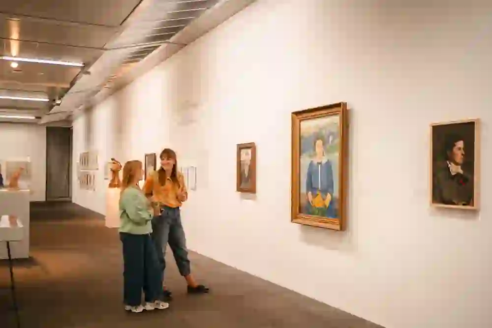 Das Bild zeigt zwei junge Frauen in einer Kunstgalerie, die sich lebhaft unterhalten. Sie stehen neben Gemälden und wirken fröhlich und entspannt, was eine Atmosphäre von Leichtigkeit und kulturellem Genuss in der Umgebung klassischer Kunst vermittelt.