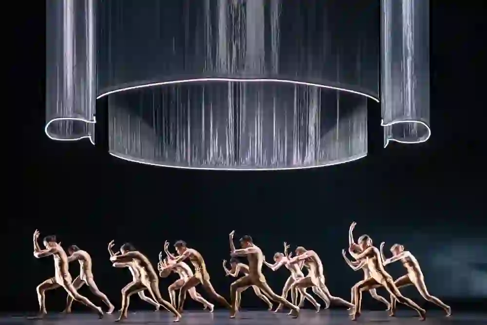 Das Bild fängt eine dynamische Ballettszene ein. Tänzerinnen und Tänzer in goldenen Kostümen bewegen sich expressiv unter einem grossen, schimmernden Kronleuchter. Ihre Pose und die Intensität des Moments vermitteln eine energiegeladene, fast elektrische Atmosphäre, die das Publikum der Bühnen Bern in ihren Bann zieht.