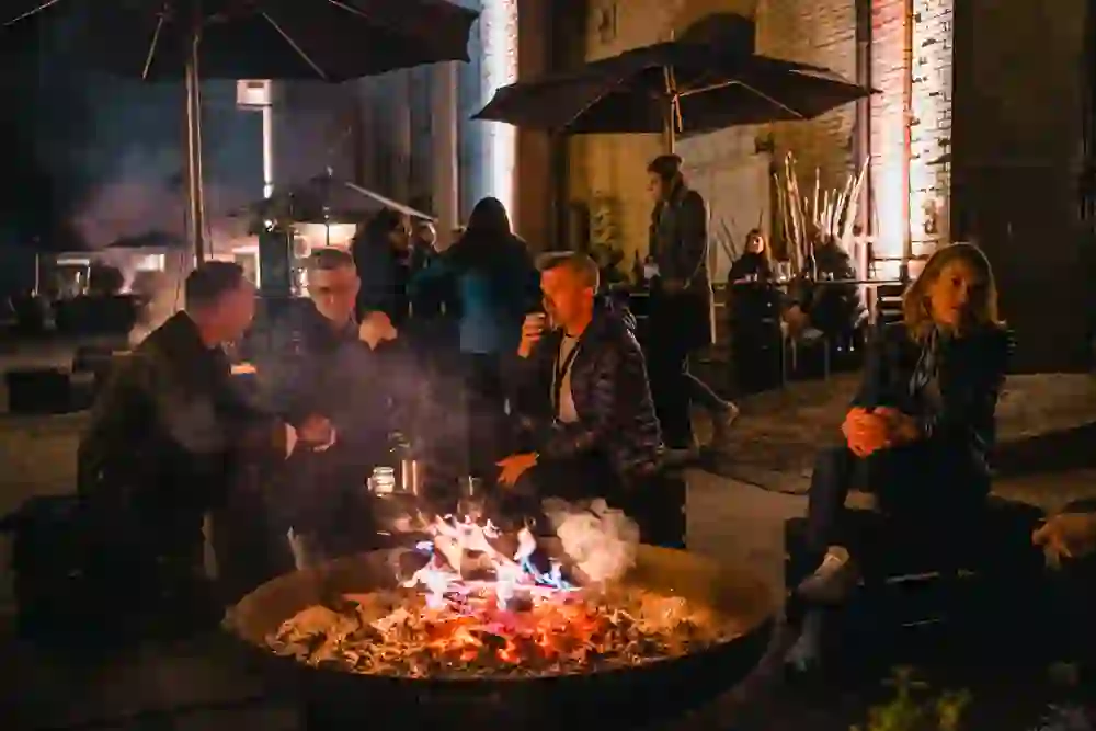 Auf dem Bild ist eine gesellige Abendrunde um ein loderndes Feuer bei Chillfood zu sehen. Personen in entspannter Atmosphäre unterhalten sich angeregt, umgeben von sanftem Licht und der Wärme des Feuers, was eine gemütliche und einladende Stimmung schafft.