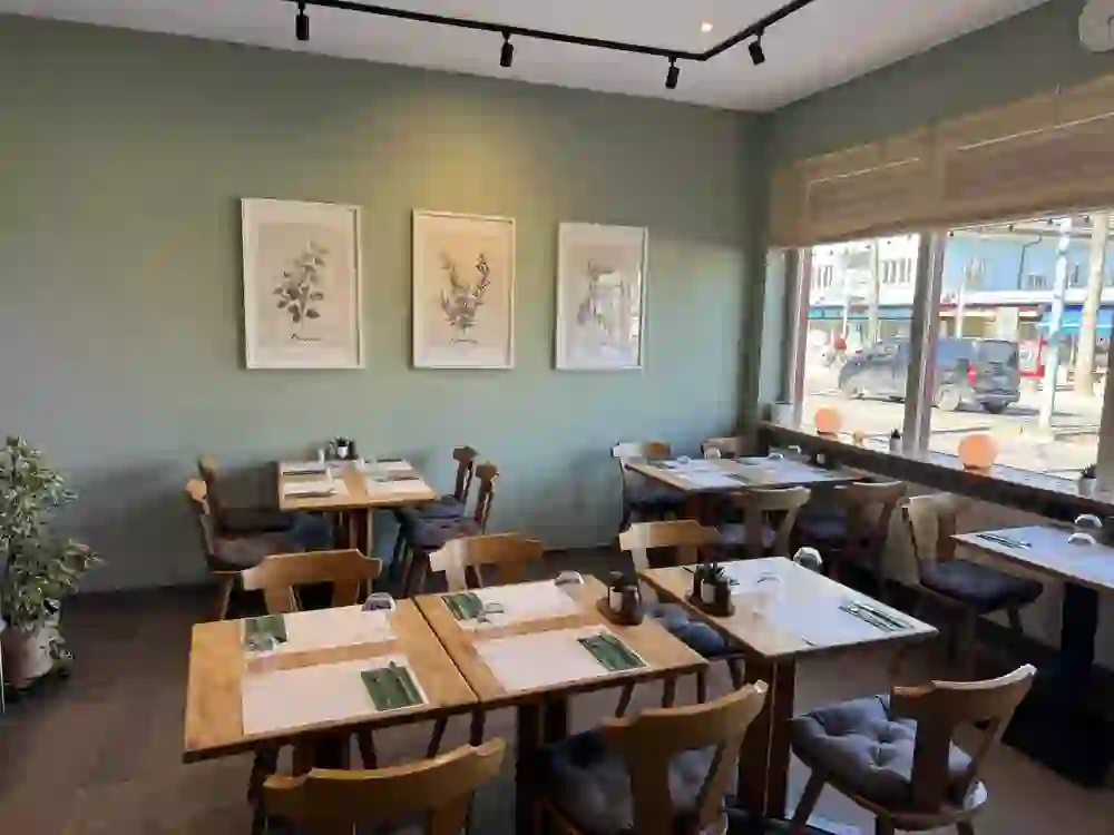 Salle de restaurant lumineuse avec des tables et des chaises en bois, joliment décorée de plantes et de tableaux botaniques. Les grandes fenêtres laissent passer une lumière chaude. L'atmosphère est calme, accueillante et détendue - un lieu où l'on se sent bien et où l'on savoure.
