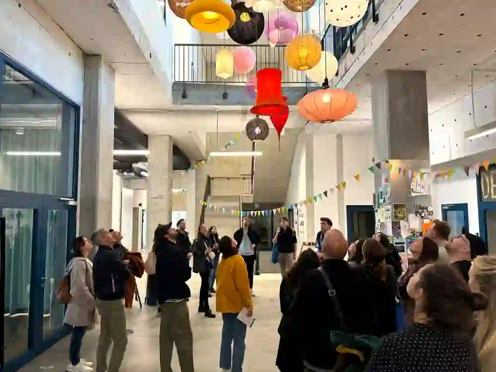 La photo montre un groupe de visiteurs* curieux lors du festival d'architecture Open House de Berne, admirant d'un œil étonné une installation de lampes créative dans un complexe de bâtiments modernes. L'atmosphère est ouverte, inspirante et pleine de joie de la découverte.