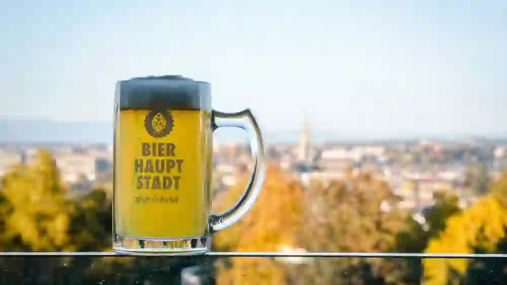 Auf dem Bild ist ein Bierkrug mit Schaumkrone zu sehen, auf dem "BIER HAUPT STADT" und "#ilovebern" steht. Der Krug steht auf einer Glasfläche mit der Berner Stadtlandschaft im Hintergrund, die herbstliche Bäume und Gebäude zeigt.