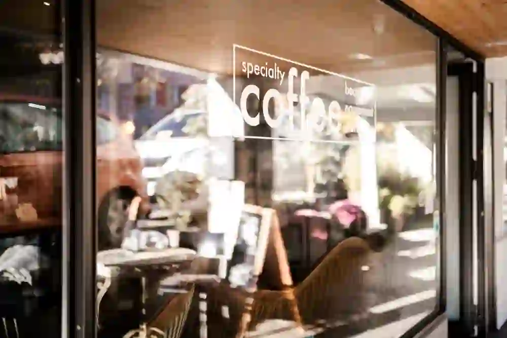 The picture shows the dreamy outdoor area of the "Coffee Coaching Club", bathed in soft sunlight that falls through a glass pane labelled "specialty coffee" and looks inviting.