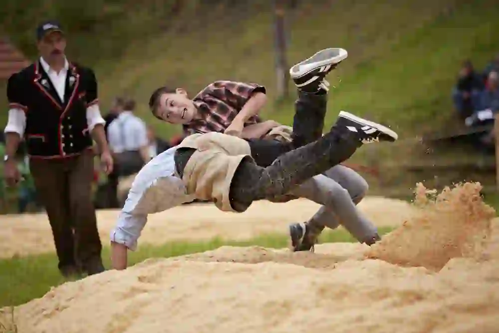 The picture shows two young men swinging in an energetic and lively scene. Their faces reflect joy and competitive spirit as sawdust sprays around them and a referee looks on attentively.