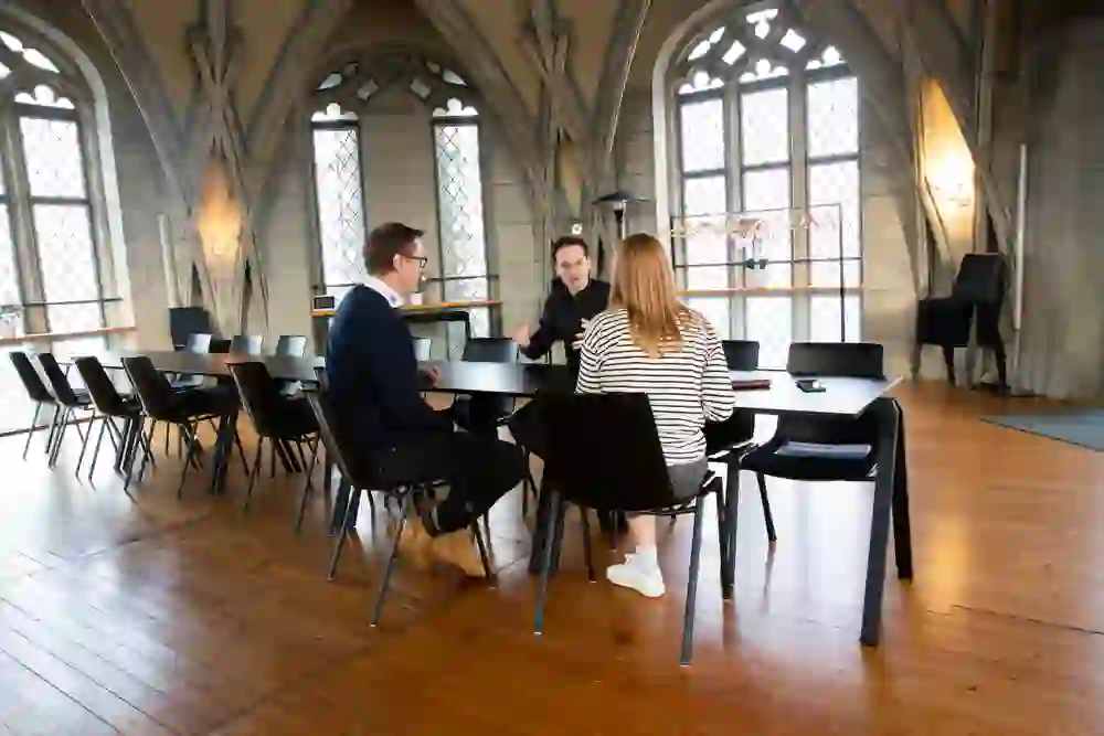 Trois personnes sont assises à une table dans une pièce au charme historique et mènent une conversation. L'ambiance semble concentrée, avec une touche d'élégance, entourée d'une architecture gothique et d'un sol en bois chaleureux.