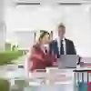 Das Bild zeigt eine professionelle, moderne Büroumgebung mit zwei Personen, die konzentriert an einem Laptop arbeiten. Die Frau im roten Blazer deutet auf den Bildschirm, während der Mann im Anzug aufmerksam zuhört. Die helle, aufgeräumte Atmosphäre vermittelt Effizienz, Zusammenarbeit und Kompetenz.