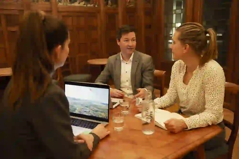 Drei Personen sitzen um einen Holztisch in einem traditionell eingerichteten Raum und führen ein Meeting, mit einem Laptop, auf dessen Bildschirm "BERN CONGRESS HUB" steht, Wasserflaschen und Dokumenten auf dem Tisch.