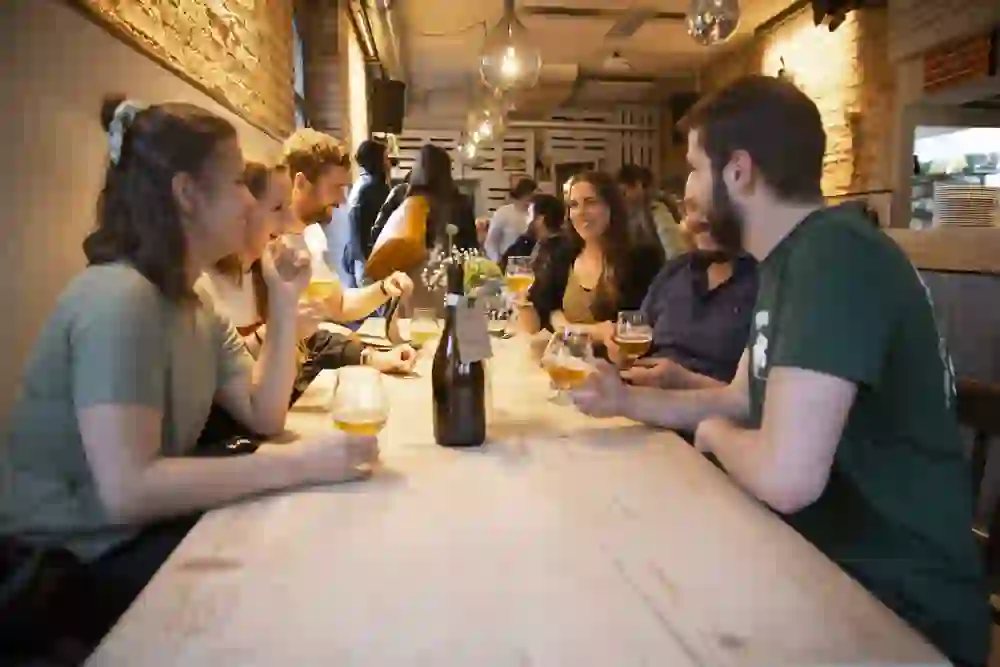 Das Foto zeigt eine fröhliche Gruppe von Menschen, die gemeinsam an einem langen Tisch in einer Bar sitzen. Sie trinken Bier, und sind in angeregte Gespräche vertieft. Die Atmosphäre ist locker und gesellig, und die warme Beleuchtung trägt zu einer gemütlichen und einladenden Stimmung bei. Die Personen lächeln und lachen, was auf eine entspannte und vergnügte Runde hindeutet.
