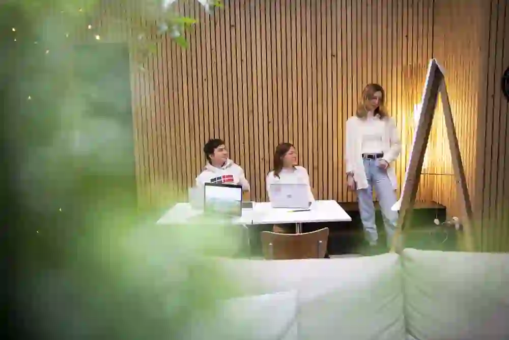 Drei Personen in einem warmen, freundlichen Raum mit Holzverkleidung und Pflanzen arbeiten zusammen; zwei sitzen am Tisch mit Laptops, während eine steht und auf eine Präsentation zeigt, wobei alle konzentriert und engagiert wirken.