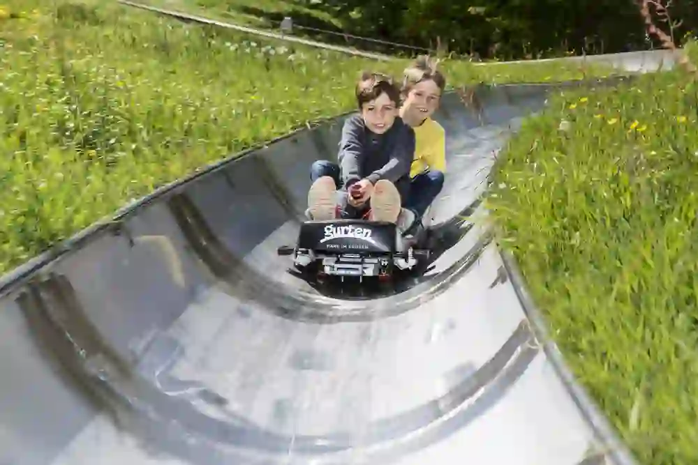 Deux enfants souriants profitent d'une descente rapide sur la piste de luge du Gurtenpark, entourés d'une nature en fleurs et d'une verdure vivante. Le plaisir et l'excitation qui se lisent sur leurs visages sont contagieux.