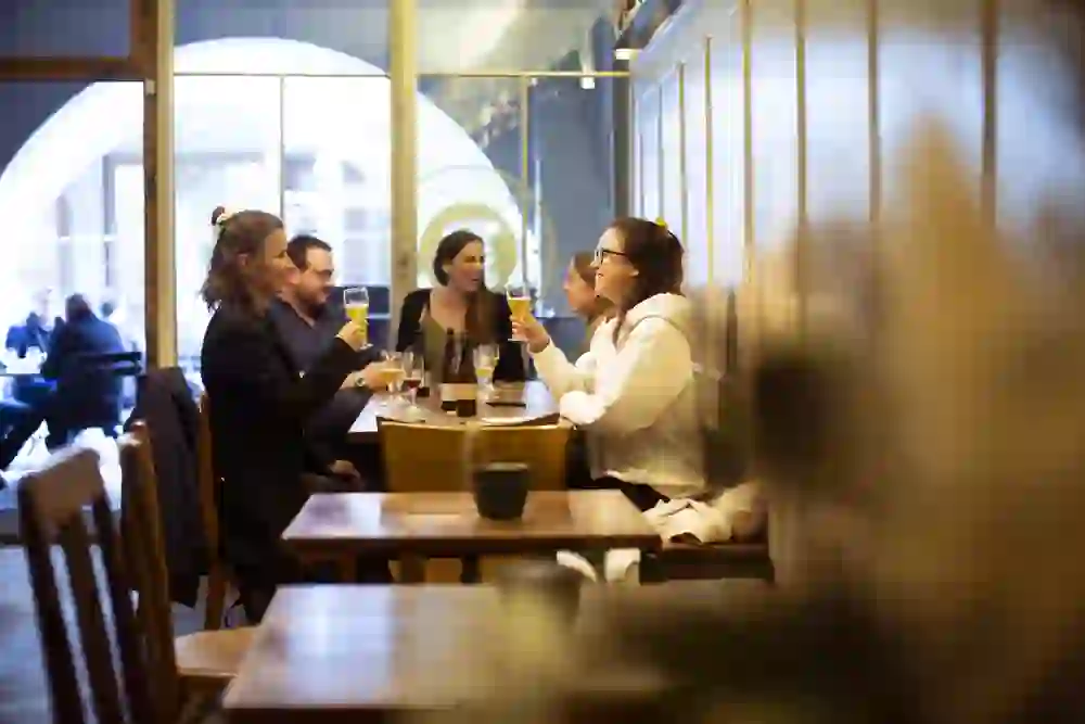 The photo shows a cheerful group of people in a bar, drinking and tasting beer together and chatting animatedly. The setting is warm and inviting, with natural light streaming through the windows creating a friendly, relaxed atmosphere as they raise a glass and enjoy each other's company.