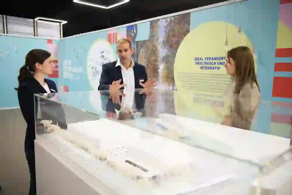 Das Bild zeigt zwei Frauen und einen Mann, die an einem Ausstellungsstand mit einem Modellgebäude und Infografiken im Hintergrund stehen und diskutieren.