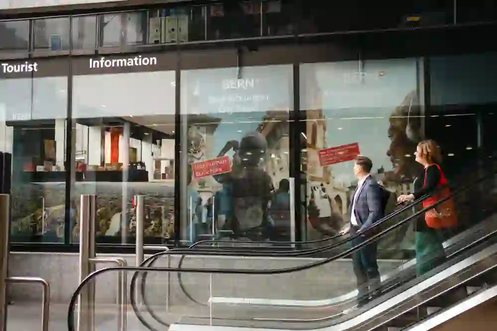 Due persone su una scala mobile salgono verso un centro di informazioni turistiche con cartelli per i tour della città, rilassati e in attesa delle esperienze che verranno.