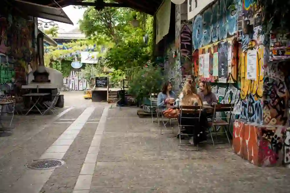 In einem kreativ gestalteten Innenhof sitzen drei Freunde entspannt an einem Tisch und genießen ihre Zeit miteinander. Umgeben von farbenfrohen Graffiti und urbaner Kunst herrscht eine lebendige, doch zugleich gemütliche Atmosphäre, die zum Verweilen einlädt.