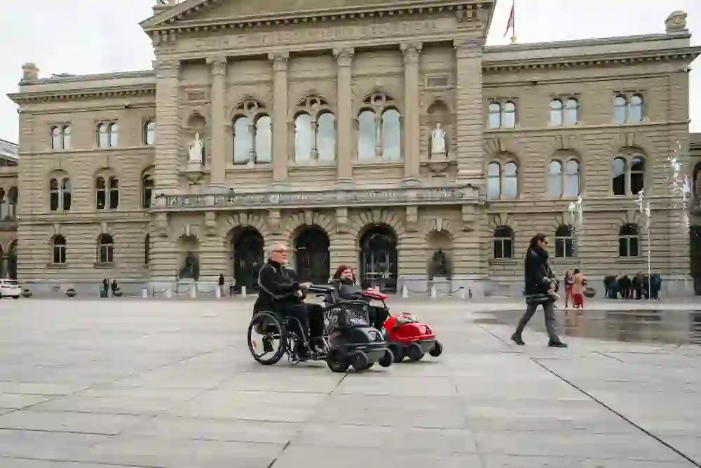 Das Bild zeigt zwei Rollstuhlfahrer auf dem Bundesplatz vor dem beeindruckenden Bundeshaus in Bern. Die Szene strahlt eine Atmosphäre von Freiheit und Inklusion aus, während die Menschen den öffentlichen Raum genießen.