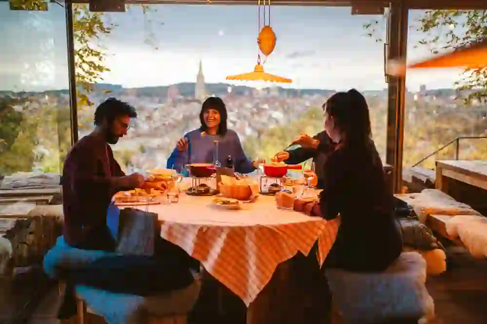 Auf dem Bild ist eine gesellige Runde im Restaurant Rosengarten in Bern zu sehen, die bei einem Fondue zusammenkommt, mit einem herrlichen Ausblick auf die Stadt Bern im Hintergrund, was eine Atmosphäre der Gemütlichkeit und Freundschaft schafft.