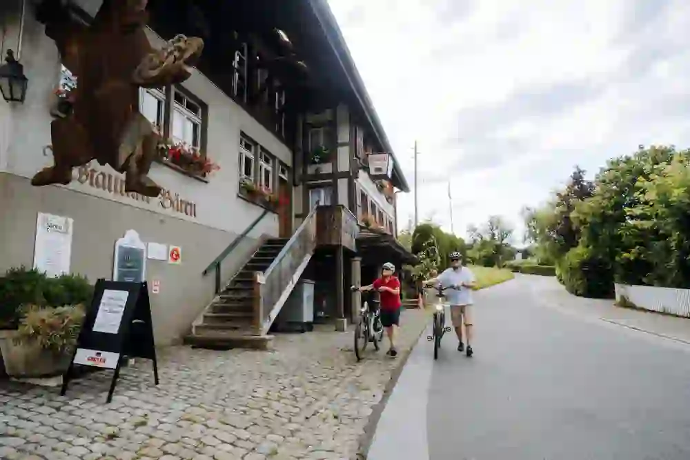 The picture shows a couple riding their bicycles in a relaxed and cheerful manner alongside a traditional Swiss inn, recognisable by its characteristic wooden façade and shutters. The scene radiates a cheerful serenity.