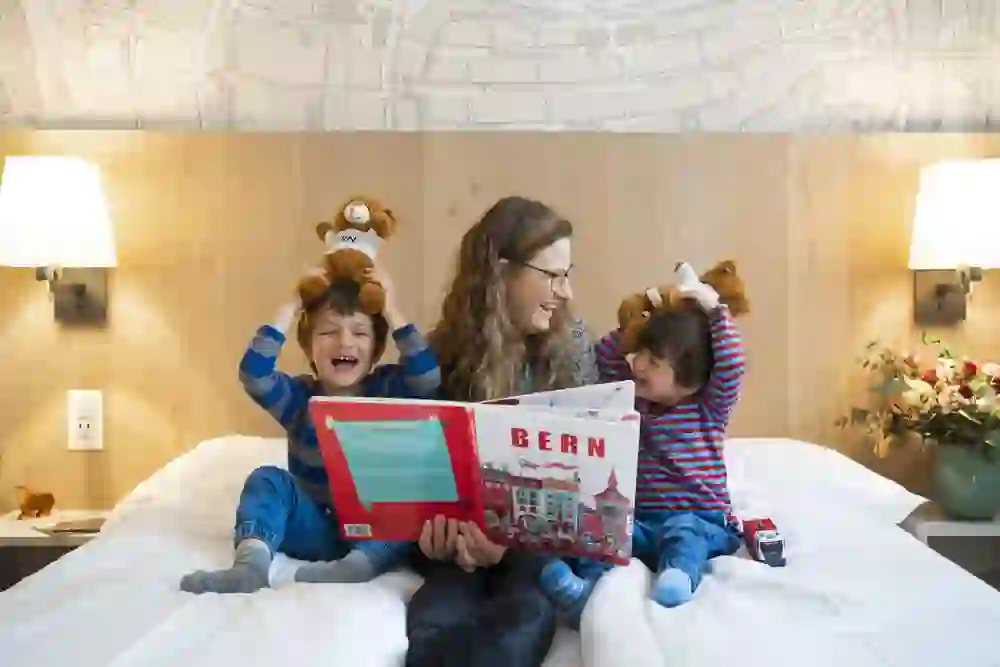 Eine Mutter liest ein Buch mit dem Titel "BERN" ihren zwei fröhlichen Kindern vor, die mit Kuscheltieren spielen, in einem gemütlichen Hotelzimmer, das Wärme und Familienglück ausstrahlt.