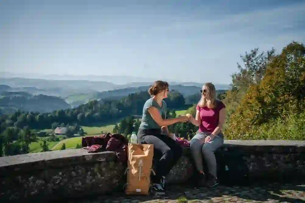 Das Foto zeigt zwei Frauen, die auf der Steinmauer bei der Lueg in Affoltern sitzend, eine Pause geniessen. Sie teilen sich einen Snack vor einer malerischen Landschaft, die sich mit saftig grünen Wiesen und Hügeln bis zu den entfernten Bergen erstreckt. Das klare Wetter und die sichtbare Freude in ihrer Interaktion spiegeln einen perfekten Tag für ein Picknick und Naturerlebnis wider.