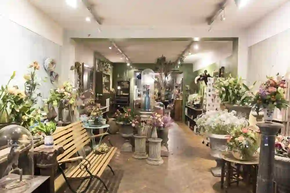 Das Bild zeigt einen Blumenladen oder eine Boutique mit einer reichen Auswahl an Blumenarrangements und Pflanzen. Die Waren sind in einem vintage-inspirierten Ambiente präsentiert, das mit rustikalen Möbeln und dekorativen Elementen ausgestattet ist.