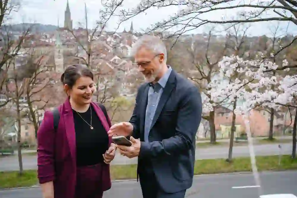 Due persone guardano insieme uno smartphone e sorridono davanti a uno sfondo pittoresco con vista sulla città e alberi in fiore, suggerendo un'atmosfera rilassata ma impegnata.