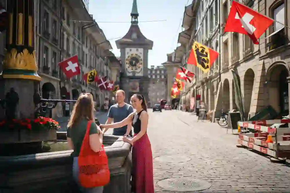 L'immagine mostra un piccolo gruppo di persone che chiacchierano davanti a una fontana in una strada di Berna. Sullo sfondo, la torre Zytglogge sovrasta la scena, circondata da bandiere svizzere e cantonali.