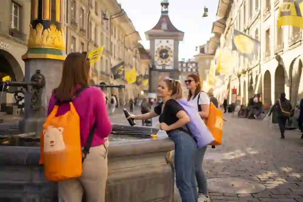Das Bild zeigt vier junge Frauen, welche mit dem Aarebag auf dem Rücken in der Stadt Bern an einem Brunnen die Flasche mit Wasser füllen