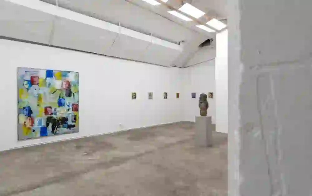 Das Bild zeigt das Innere der da mihi Galerie im Kunstkeller mit einem großen abstrakten Gemälde an der Hauptwand und einer Reihe kleinerer Porträts daneben. In der Mitte des Raumes steht auf einem Sockel ein skulpturales Kunstwerk. Der Raum hat ein industrielles Flair mit sichtbaren Deckenstrahlern und einer Betonstruktur, die eine moderne und minimalistische Stimmung erzeugt.