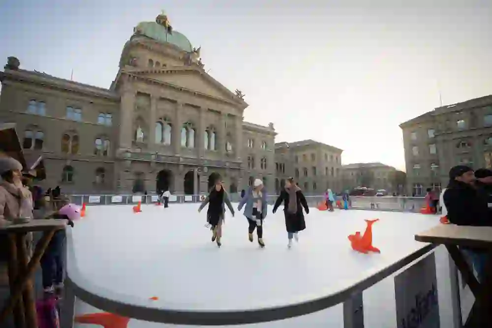 Das Bild zeigt drei Frauen, die an der Kunsteisbahn auf dem Bundesplatz in Bern stehen und anstoßen. Im Hintergrund ist das Bundeshaus zu sehen. Die Frauen tragen Winterkleidung und lachen miteinander. Auf der Eisbahn laufen Kinder Schlittschuh, einige benutzen orangefarbene Lernhilfen. Die Atmosphäre wirkt winterlich und fröhlich, und die Szenerie wird von der untergehenden Sonne beleuchtet, was eine warme und gemütliche Stimmung erzeugt.