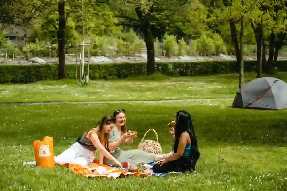 The picture shows three women having a picnic on the green meadow at the Eichholz campsite in Bern. They are sitting on colourful bath towels, eating and chatting happily. The River Aare, trees, bushes and a tent can be seen in the background. The atmosphere is relaxed and summery.