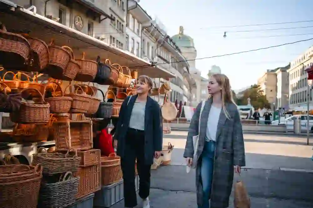 Zwei Frauen betrachten neugierig einen Stand mit Körben auf einem belebten Markt, umgeben von städtischem Treiben und historischer Architektur. Die Atmosphäre ist aufgeschlossen und lebhaft.