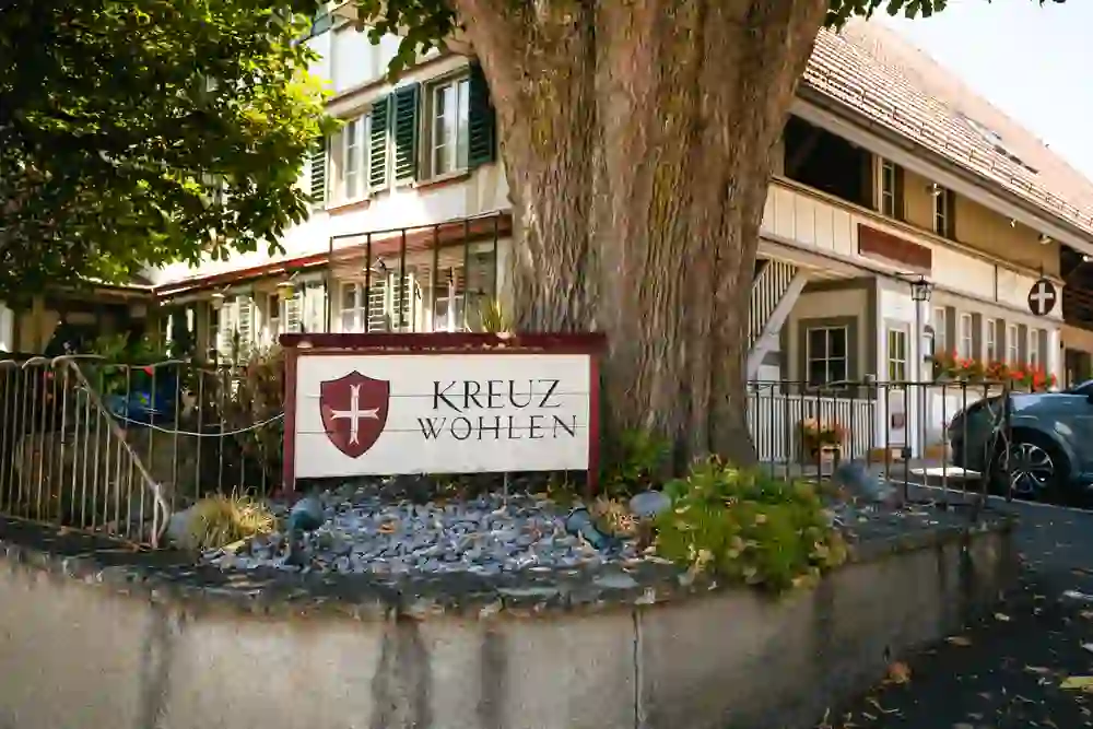 The picture shows the entrance area of the "Kreuz Wohlen" inn. In the foreground is a large sign with the coat of arms and the inscription "Kreuz Wohlen". The sign is in front of a large tree and is surrounded by a small garden with plants. The inn can be seen in the background, a traditional building with green and white shutters and flowers in the windows. Part of a parked car is also visible. The scene is inviting and idyllic.
