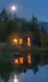 Small wooden house by a quiet pond, warmly lit under the clear moonlit sky, surrounded by forest and mountains. The reflection in the water enhances the peaceful, almost magical atmosphere - a place full of peace, security and quiet romance.