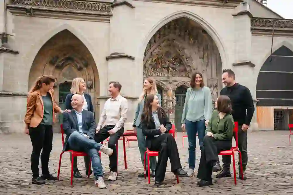Sieben lächelnde Personen posieren lässig auf roten Stühlen vor dem Berner Münster, eine gelöste und freundliche Stimmung ausstrahlend, als Gruppe vereint.
