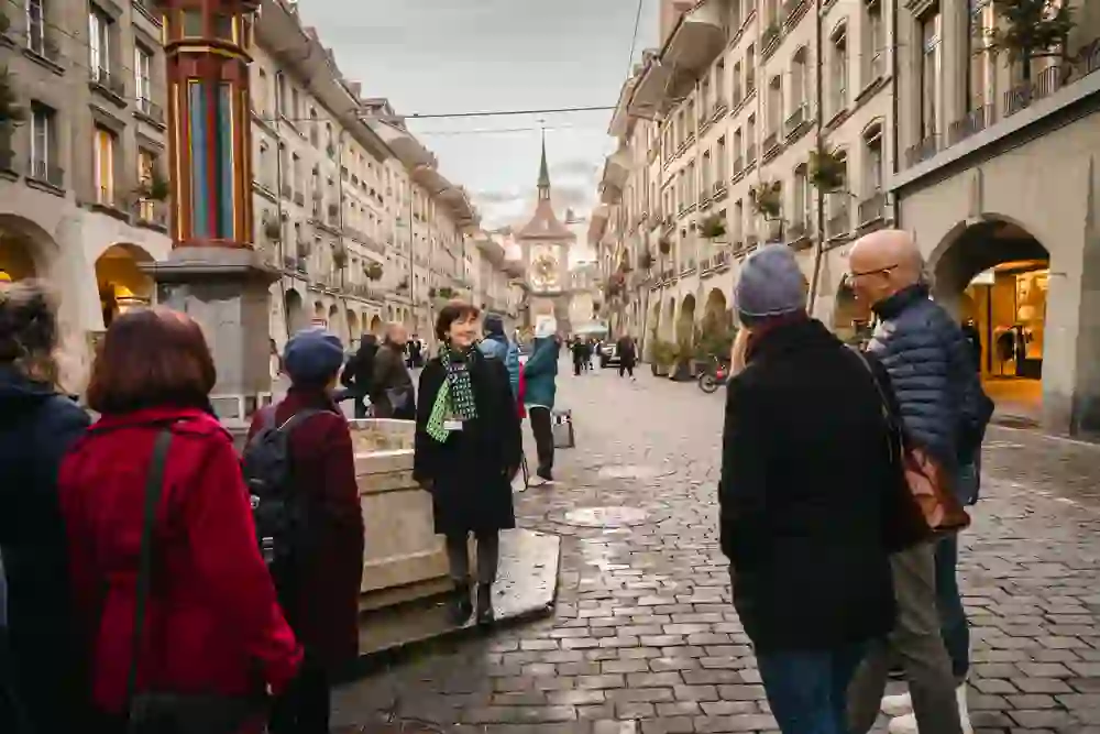 Das Foto zeigt eine Gruppe von Menschen, die an einer Stadtführung teilnehmen. Sie stehen in einer historischen Strasse in Bern mit Kopfsteinpflaster und traditionellen Gebäuden mit Arkaden. Am Ende der Strasse ist der Zytglogge mit dem Uhrwerk zu erkennen. Die Stimmung wirkt entspannt und die Teilnehmer interessiert, während sie die Erklärungen aufnehmen und die charmante Umgebung geniessen.