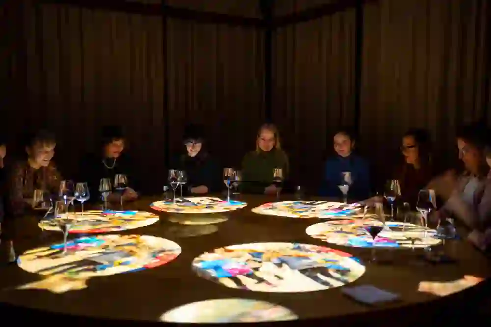 La photo montre un groupe de personnes réunies autour d'une table éclairée au casino de Berne. La lumière projette des reflets colorés sur la surface de la table, ce qui crée une atmosphère intime et conviviale.