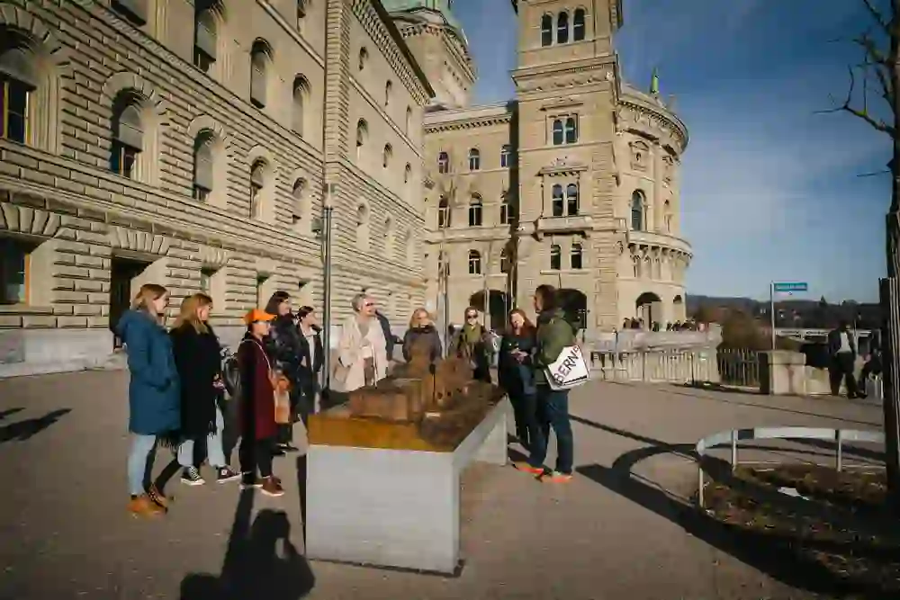 Eine Gruppe von Zuhörern steht vor dem Bundeshaus in Bern und lauscht einem Führer. Sie blicken auf ein Modell des Gebäudes, ihre Gesichter von der Sonne beschienen, was auf eine aufschlussreiche und angenehme Führung „1848 – Bern wird Bundesstadt“ hindeutet.
