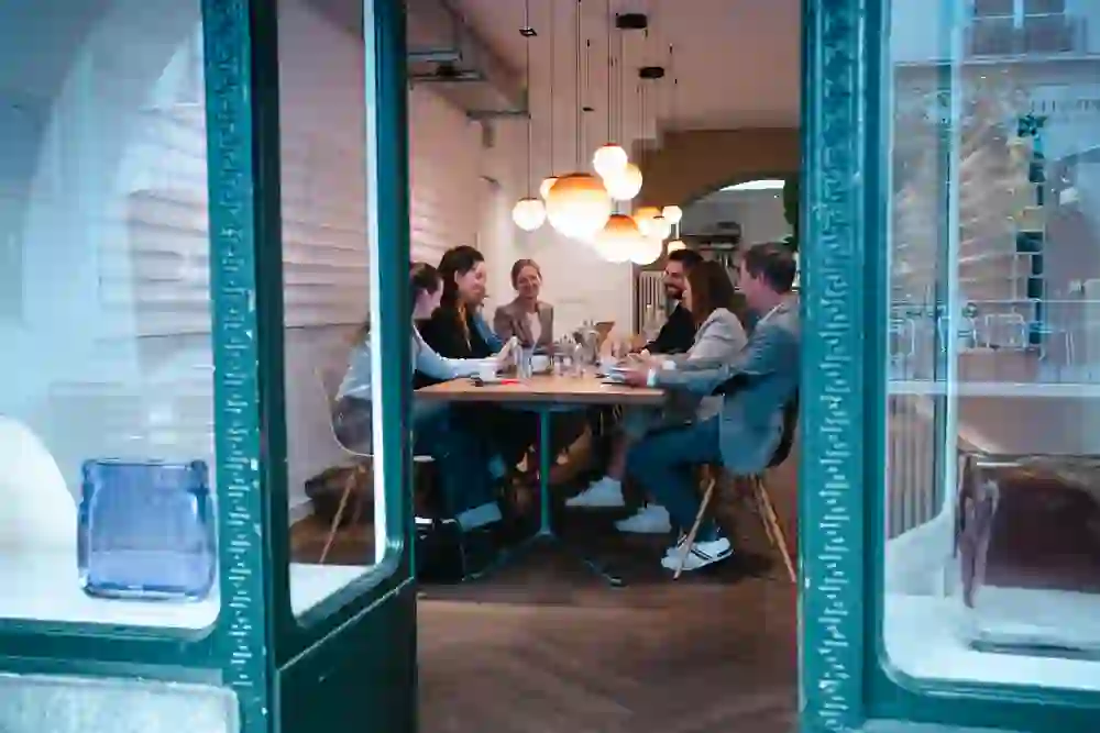 Das Bild zeigt einen Blick durch ein Fenster auf eine Gruppe von Menschen, die an einem Tisch in einem gemütlich beleuchteten Raum sitzen und zu interagieren scheinen, möglicherweise bei einem Meeting oder geselligen Beisammensein.