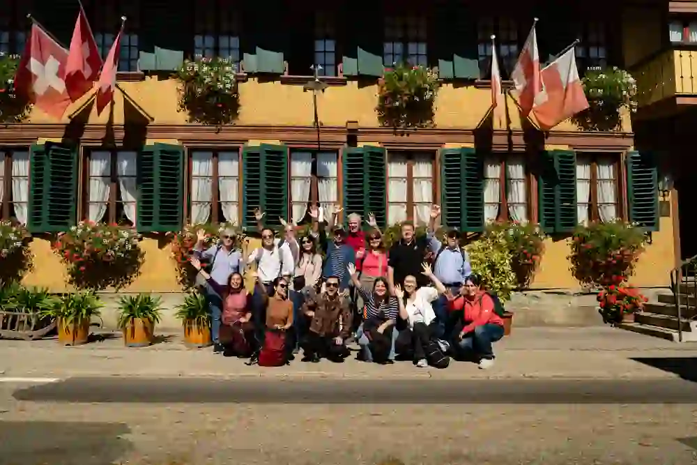 Eine fröhliche Gruppe posiert vor einem charmanten, blumengeschmückten Haus mit Schweizer Flaggen. Die ausgelassene Stimmung und das sonnige Wetter spiegeln pure Lebensfreude und Gemeinschaft wider – ein Moment voller Herzlichkeit und Zusammenhalt.