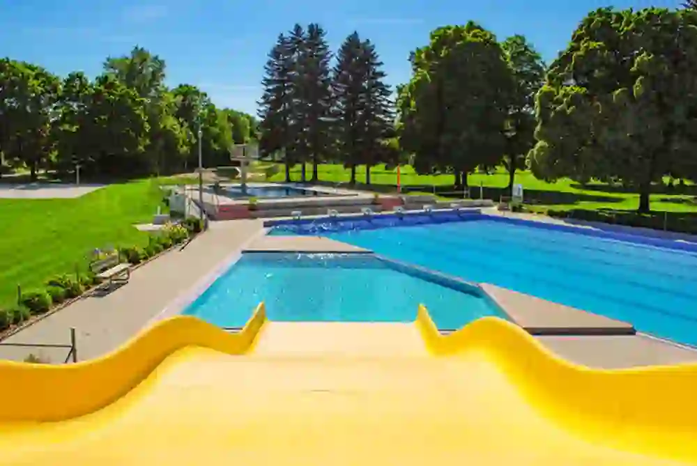 Das Bild fängt eine idyllische Szene in einem Freibad ein, umgeben von üppigen Bäumen unter klarem blauem Himmel. Die Sonnenstrahlen und das ruhige, einladende Wasser schaffen eine entspannende und erfrischende Atmosphäre, perfekt für einen Sommertag.