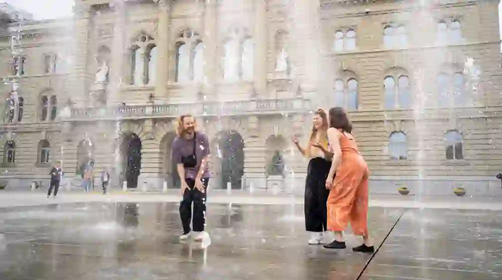Vor der historischen Kulisse des Bundesplatzes in Bern vergnügen sich zwei Frauen und ein Mann im spritzenden Wasserspiel, ihre Gesichter von Freude erfüllt. Es ist ein lebendiger, fröhlicher Moment inmitten der Stadt.
