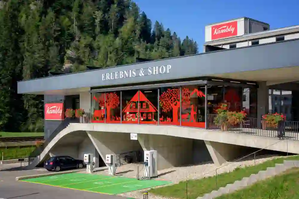 La photo montre l'Aventure & Boutique Kambly, nichée dans un cadre idyllique et verdoyant. Une ambiance lumineuse et estivale entoure le bâtiment moderne avec ses éléments décoratifs rouges distinctifs qui reflètent le design suisse traditionnel. Les stations de recharge électrique à l'avant signalent la durabilité.