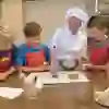 Une joyeuse chef en tenue de cuisine blanche dirige trois garçons concentrés lors de l'événement pâtisserie de Kambly. Ils observent avec attention comment le chocolat est coulé, entourés d'ustensiles de cuisson et avec un chef-d'œuvre de biscuits sur la table.