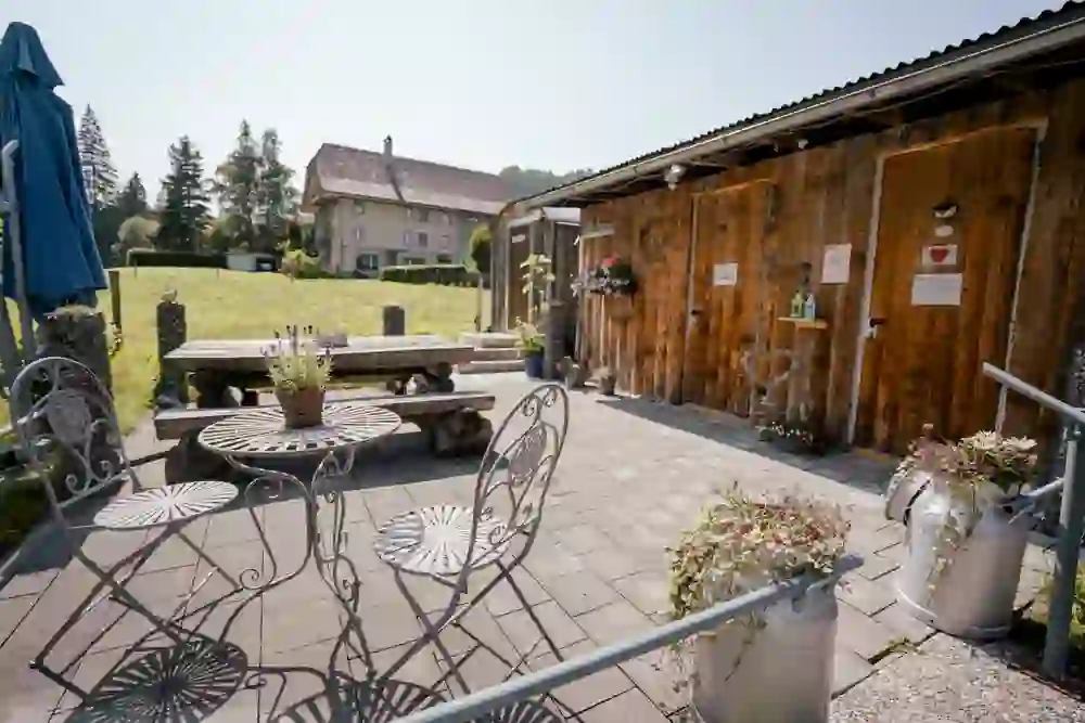 Das Foto zeigt eine idyllische Szene im Freien auf dem Hofladen Mühlihof in Rüschegg. Davor befindet sich eine gemütliche Sitzecke mit schmiedeeisernen Stühlen und einem Tisch, liebevoll dekoriert mit lila Blumen. Die warme Ausstrahlung des Holzes und die charmanten Blumentöpfe erzeugen eine einladende Atmosphäre. Alles deutet darauf hin, dass dies ein Ort ist, an dem man lokale Produkte geniessen und die Ruhe des Landlebens in vollen Zügen schätzen kann.
